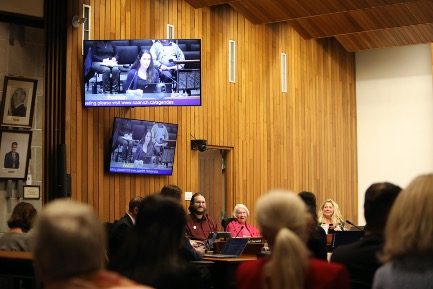 People sit in a large room for a formal meeting. Two screens on the wall show a speaker at the microphone. In the background, people at a board table with microphones are in focus. In the foreground, the backs of audience members' heads are shown.